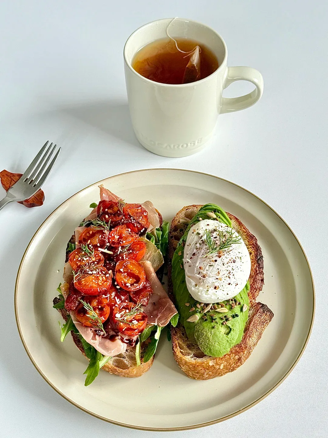 Two open-faced bread slices topped with tomatoes and avocado with an egg, beside a cup of tea.